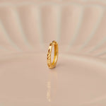 Side angle view of a slim textured 18K gold hoop nose ring placed on a soft beige surface with gentle curved light patterns