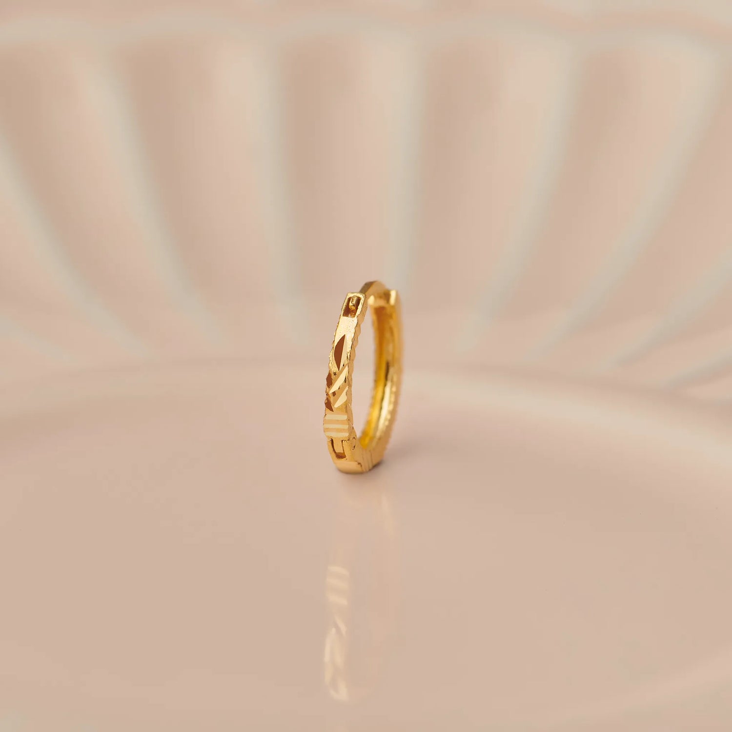 Side angle view of a slim textured 18K gold hoop nose ring placed on a soft beige surface with gentle curved light patterns