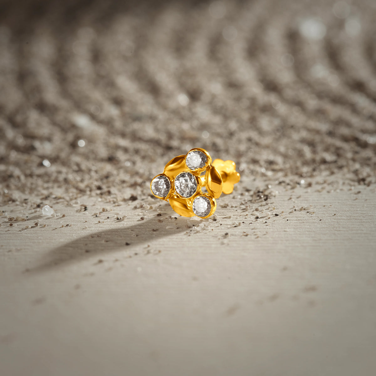 An 18K gold nose pin shaped like a flower cluster, set with clear crystal stones, photographed on a light textured surface