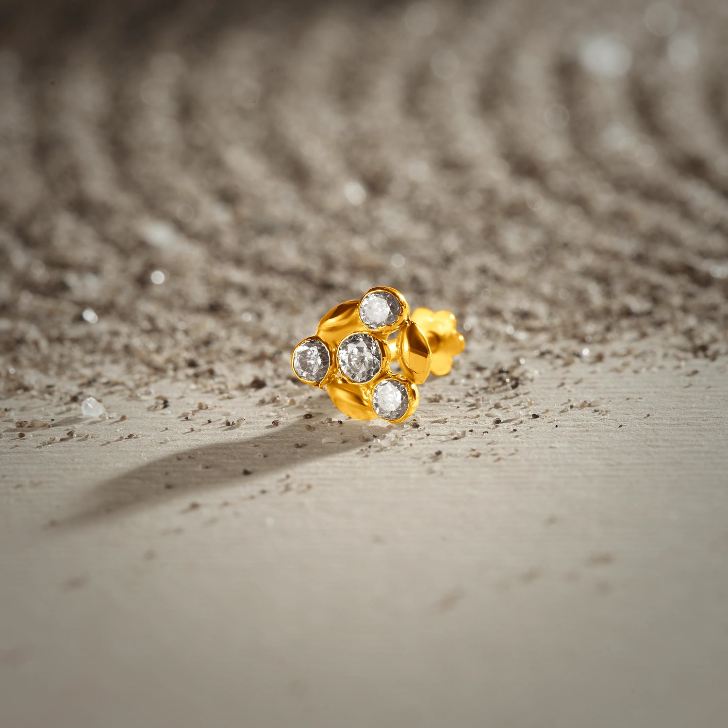 An 18K gold nose pin shaped like a flower cluster, set with clear crystal stones, photographed on a light textured surface
