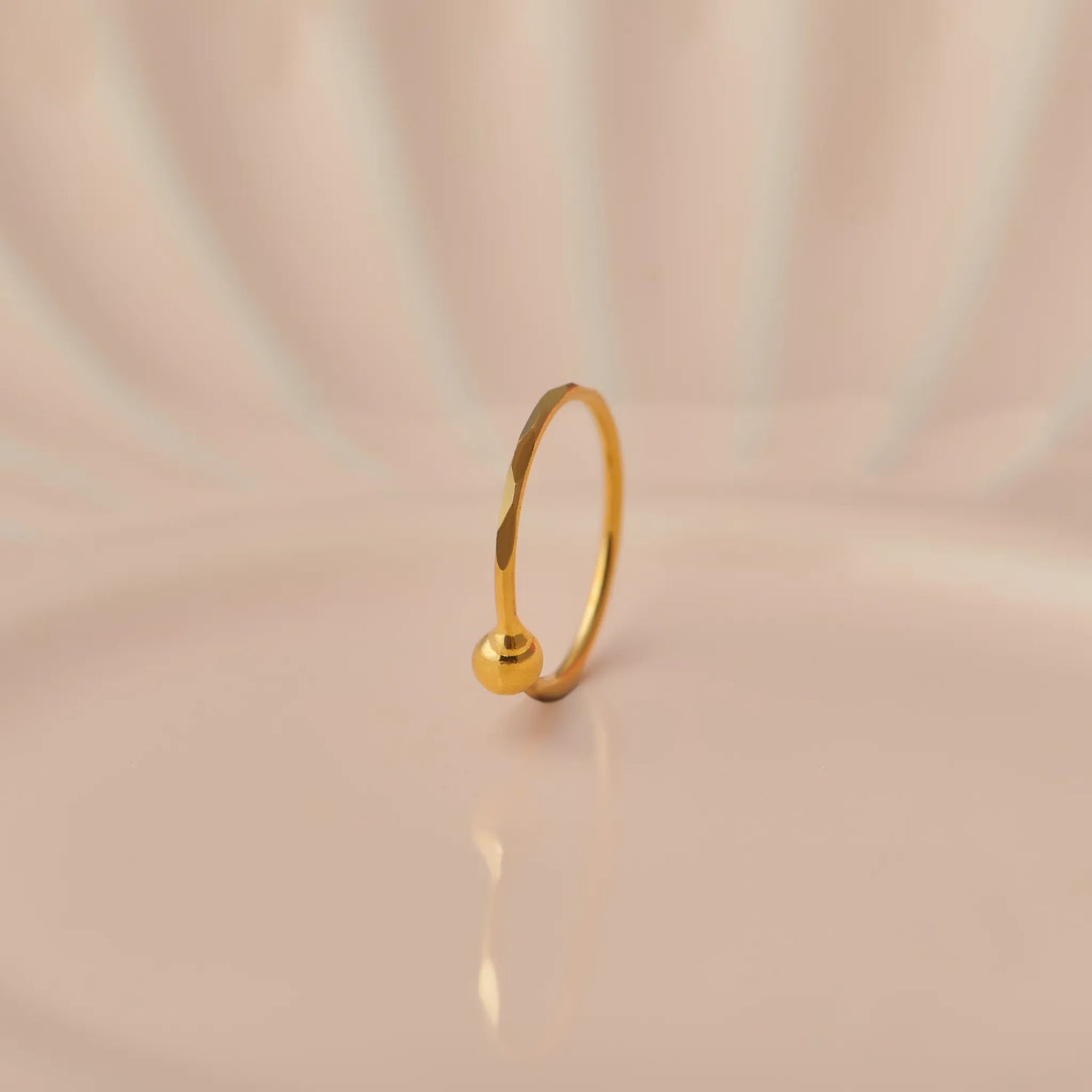 Close up of minimalist 18K gold nose ring with a small ball accent standing upright on a soft beige background with subtle light reflections