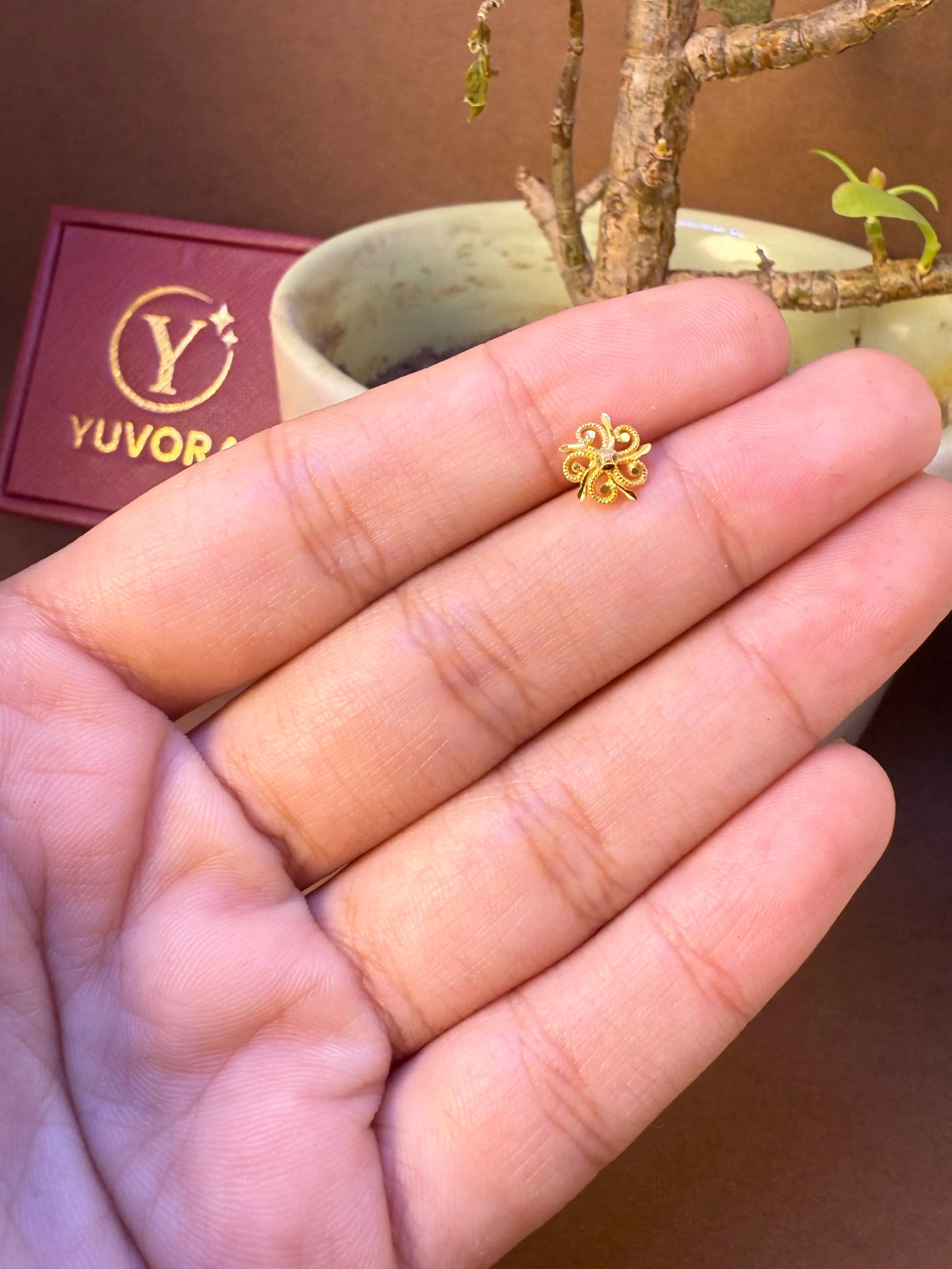 Four-petal gold nose pin with curled wire design held in hand, Yuvora jewelry box in background