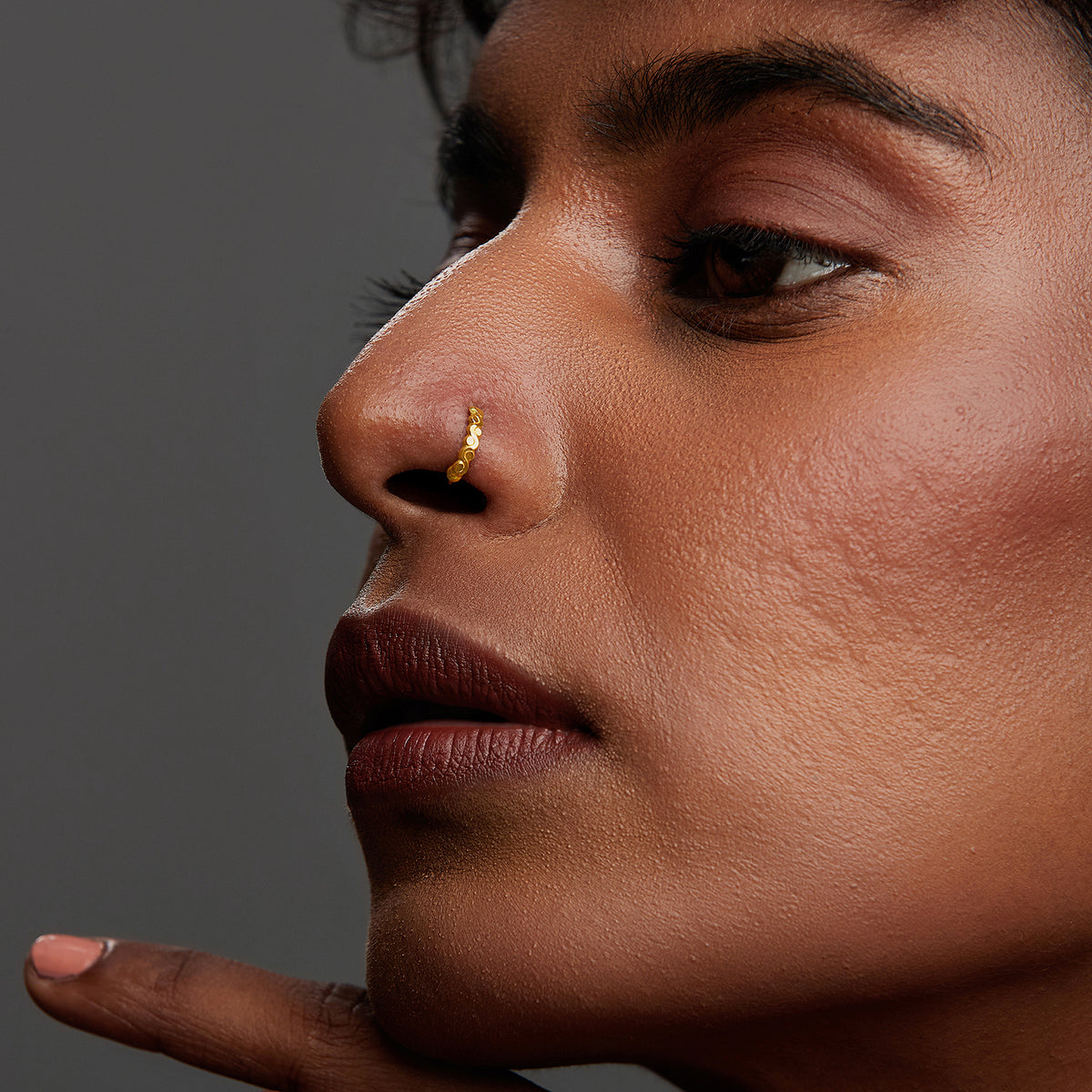 Close-up of a woman wearing a thin gold beaded hoop nose ring, hand near chin