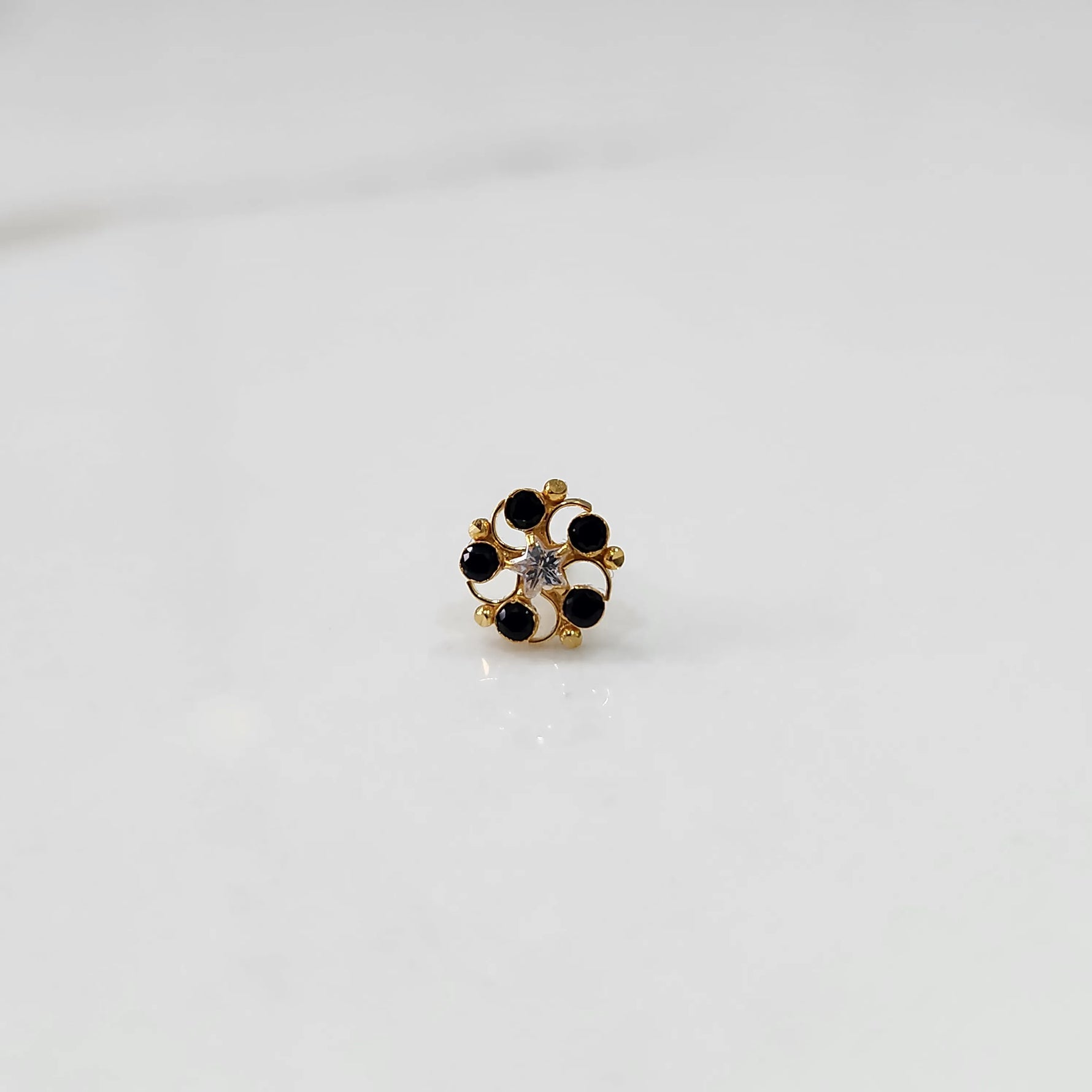 Front view of gold nose pin with black gemstone petals and clear center stone on white background