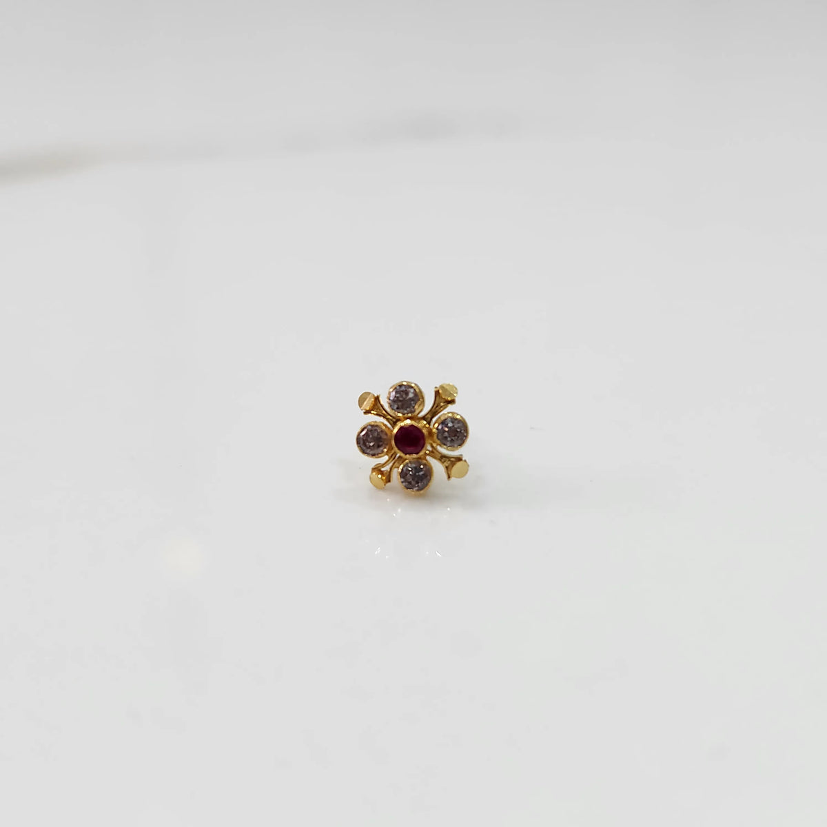 Gold nose pin with clear gemstone petals and red center stone, angled view on white background