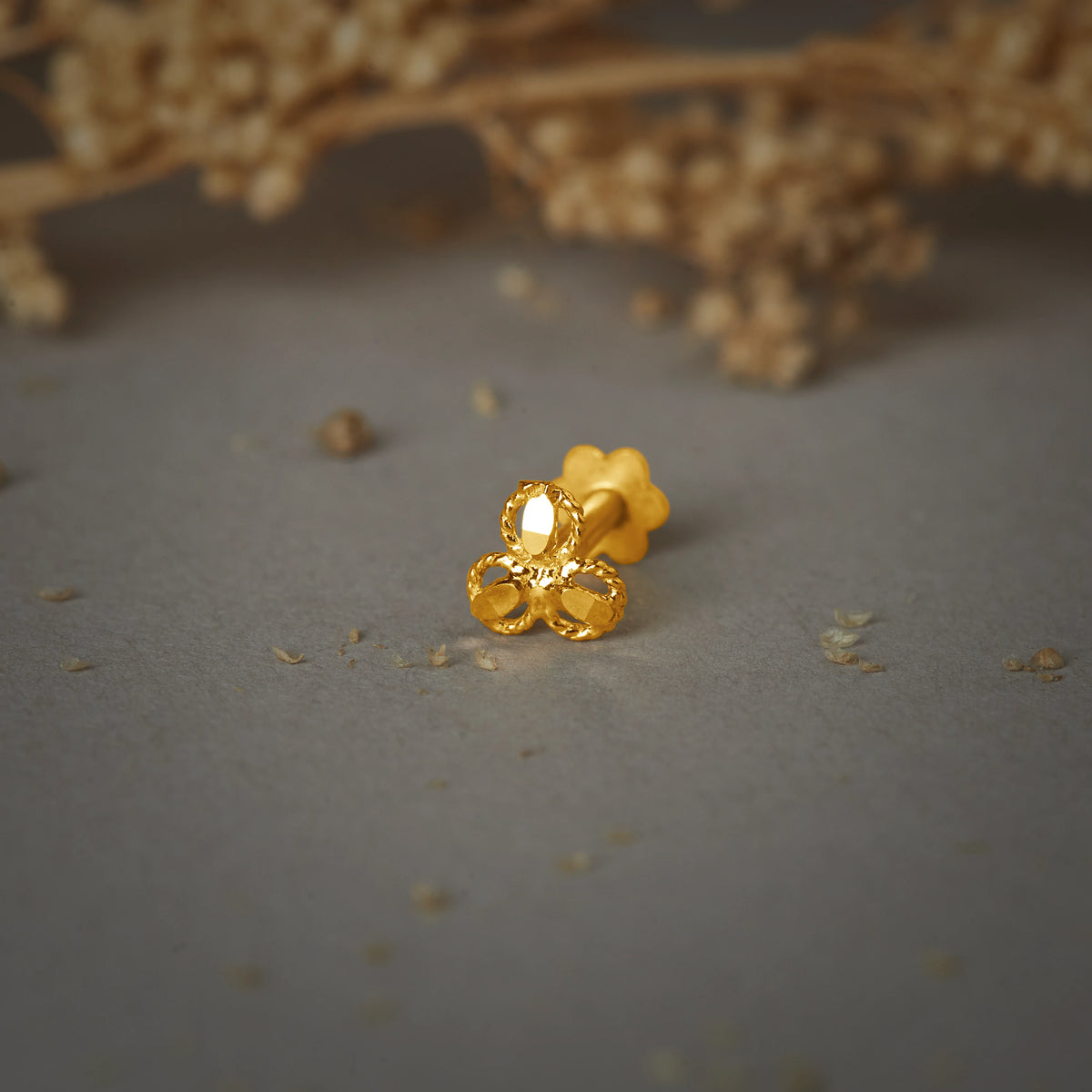 A gold clover-shaped nose pin with a textured oval top and two rounded petals below, placed on a grey textured background