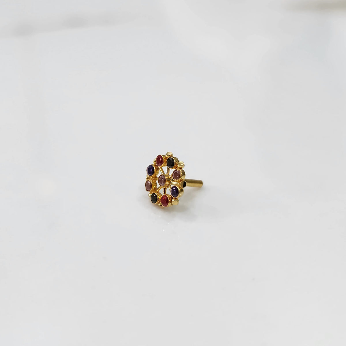 A gold cluster-design nose pin set with multiple oval multicolor stones in purple, red and pink, placed on a white background