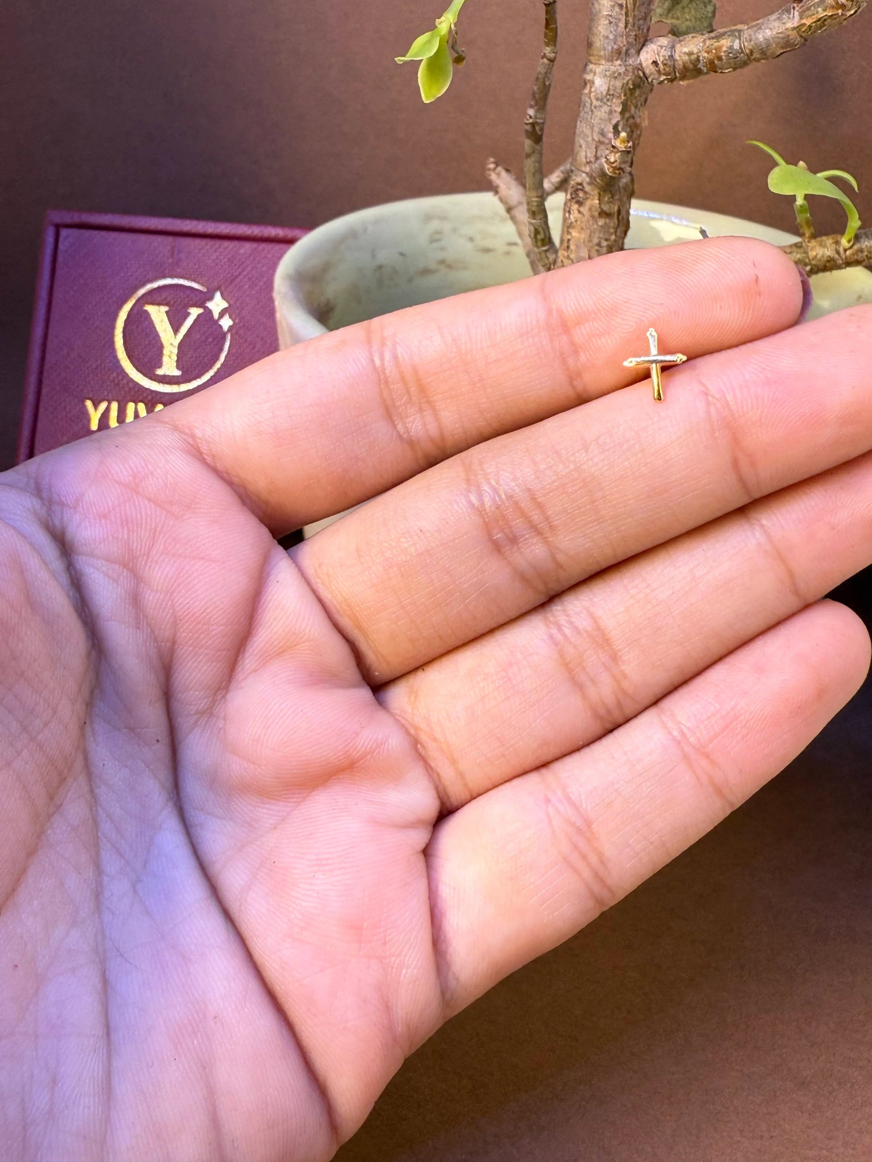 Simple cross-shaped gold nose pin held in hand, Yuvora jewelry box in background