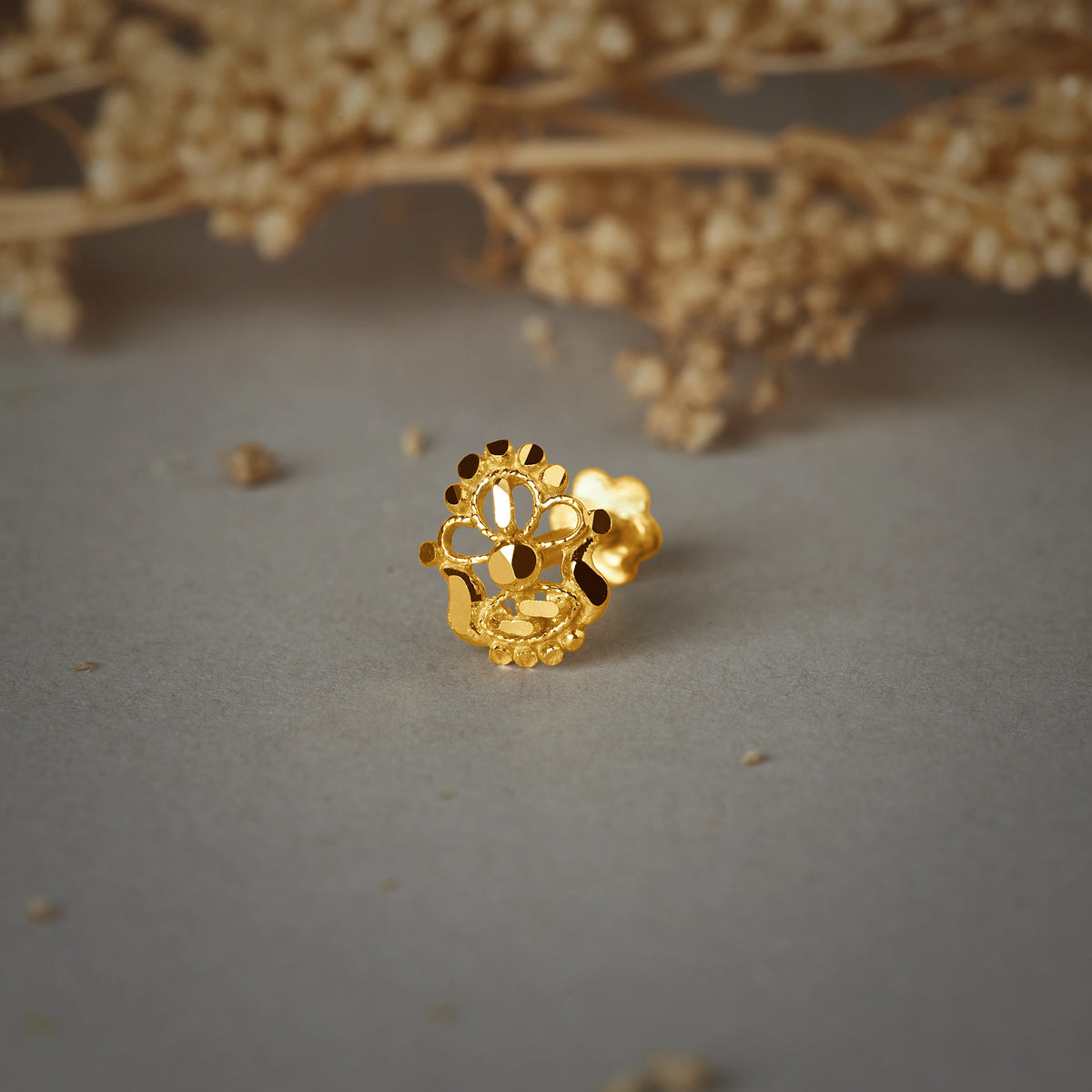A gold cutwork nose pin with a floral open design and a beaded lower edge, placed on a grey background with dried flowers