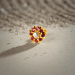 A gold eight-petal flower stud earring with ruby stones around a white center stone, placed on a textured beige fabric background