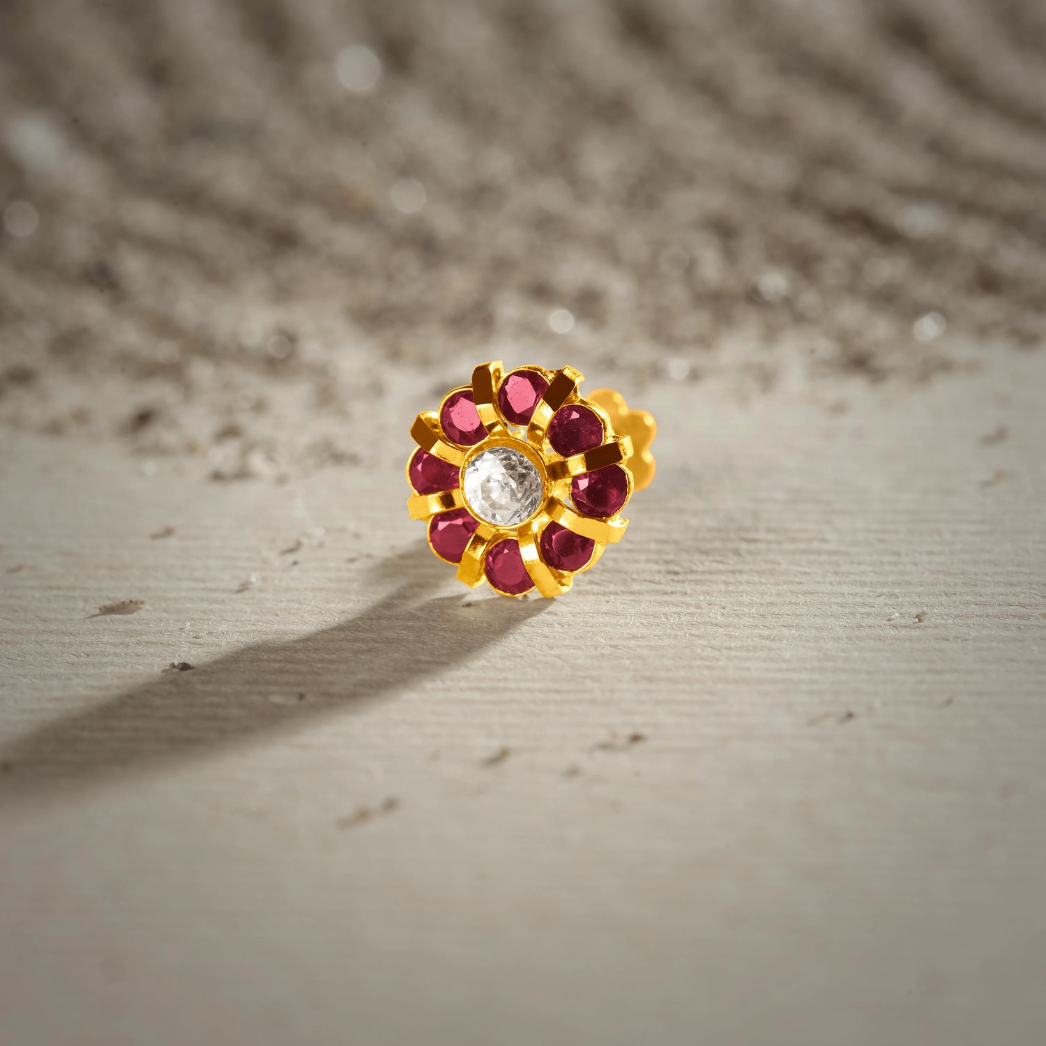 A gold eight-petal flower stud earring with ruby stones around a white center stone, placed on a textured beige fabric background