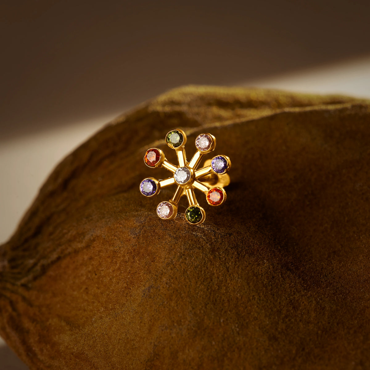 A gold eight-ray stud earring with a white center stone and multicolor stones at each ray tip, placed on a textured brown fabric background