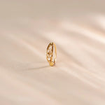 Close up of an 18K gold nose ring with intricate cutwork detailing standing upright on a soft beige fabric background with gentle light reflections