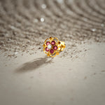 A gold floral stud earring with ruby stones and a white center stone, placed on a textured beige fabric background