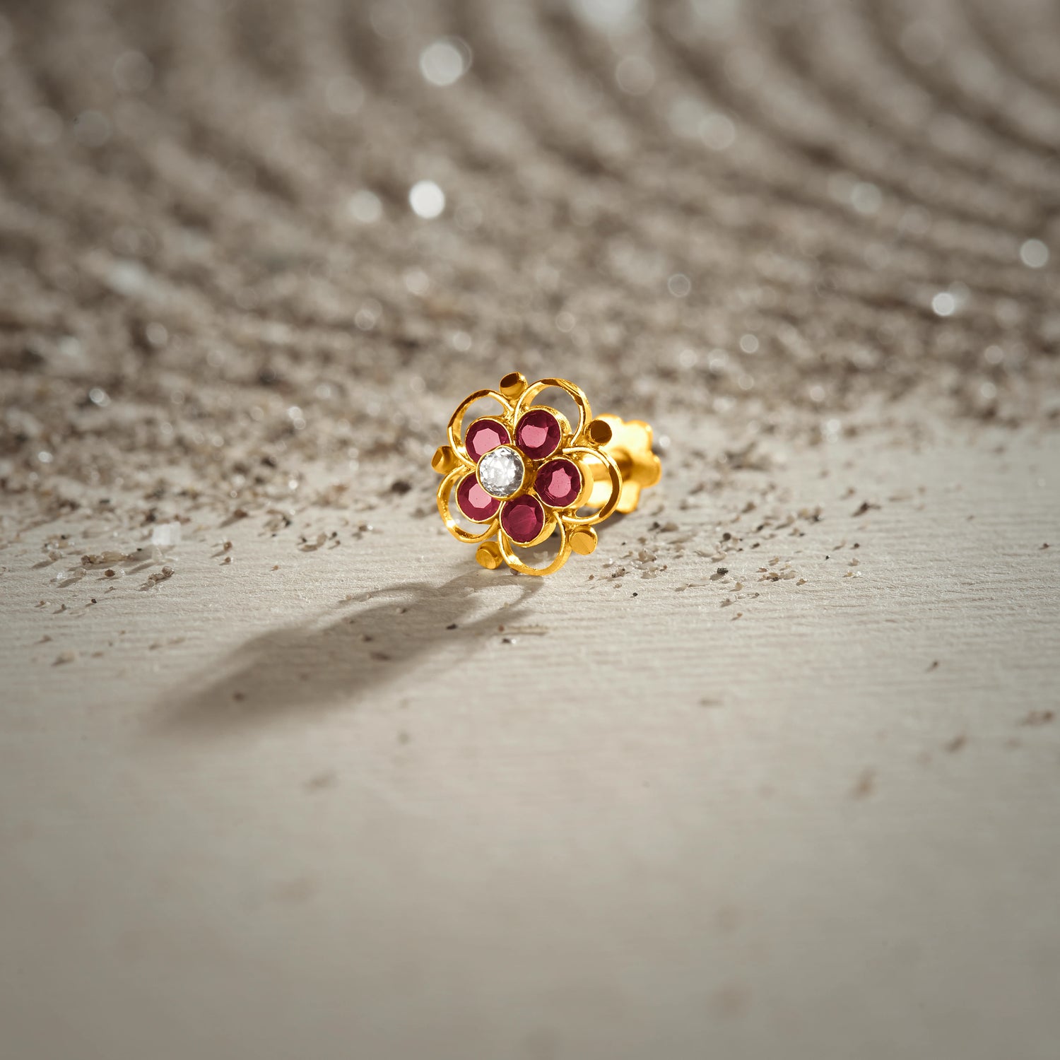 A gold floral stud earring with ruby stones and a white center stone, placed on a textured beige fabric background