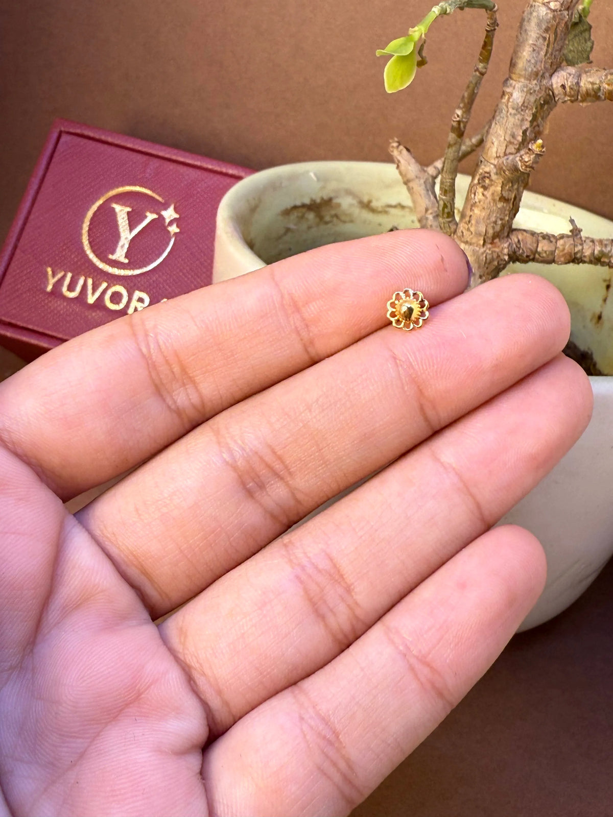 Small gold flower nose pin held on fingers for size comparison, with Yuvora jewellery box and plant in background