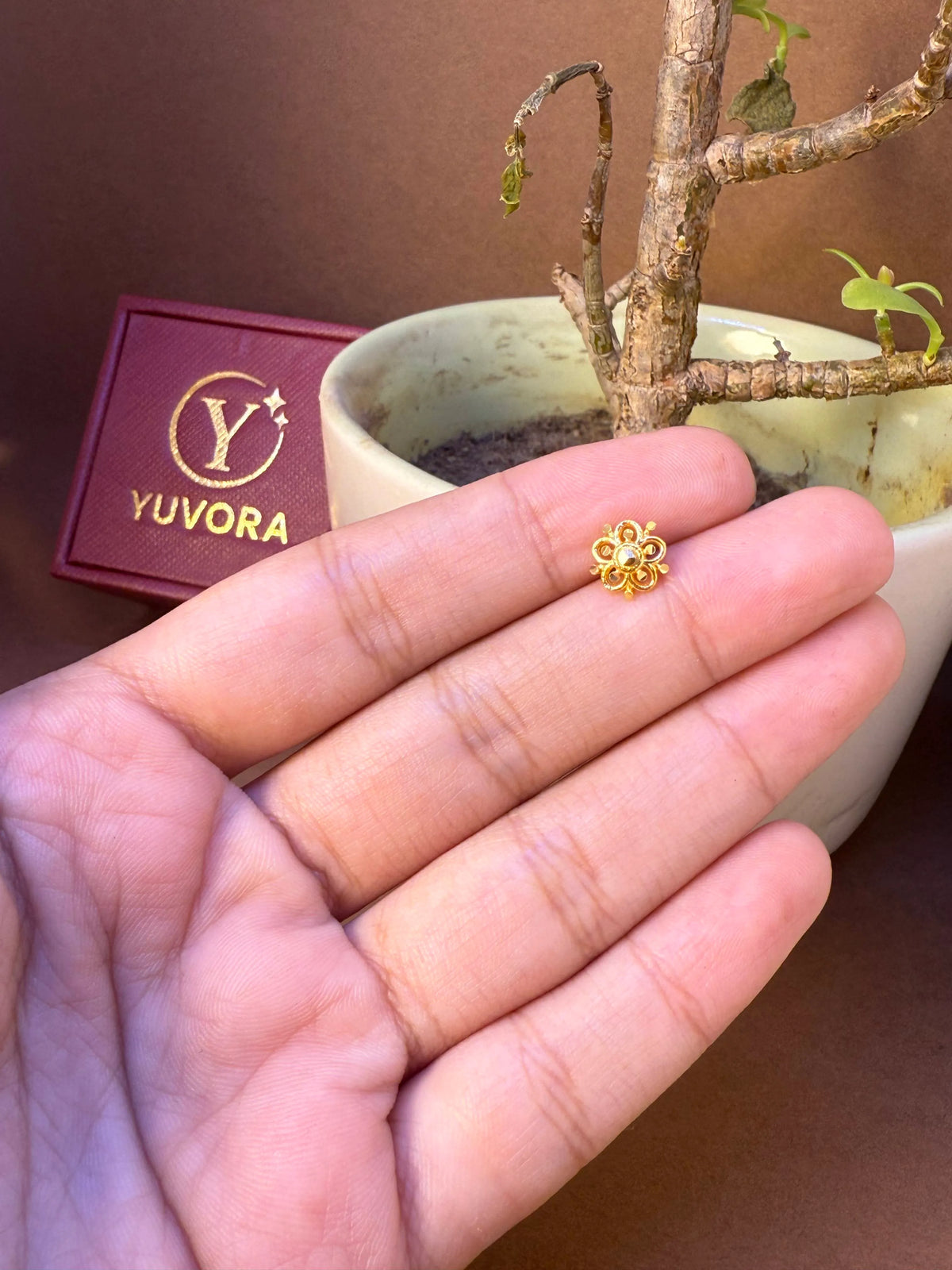 Small flower-shaped gold nose pin held in hand, with Yuvora jewelry brand packaging in the background