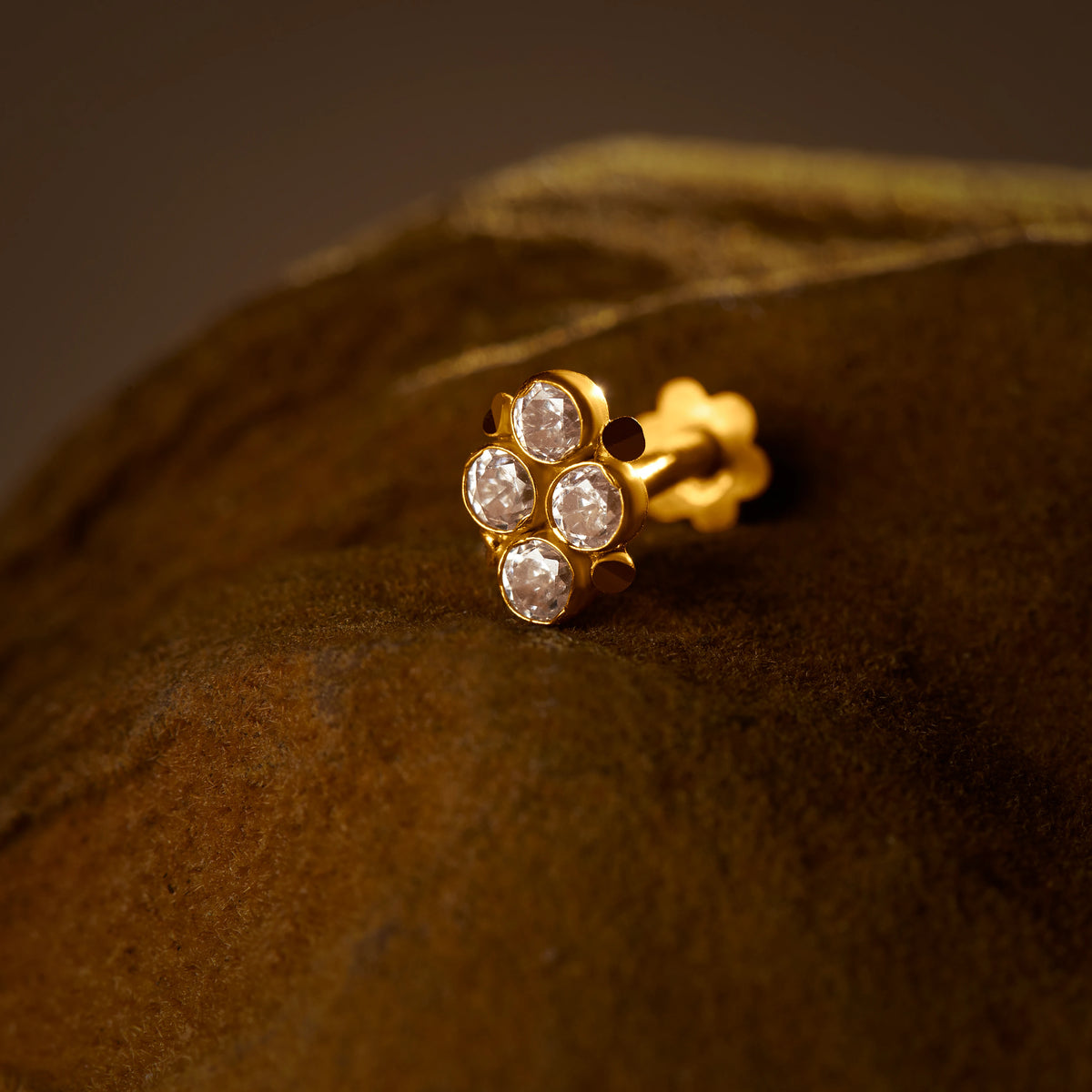 Women's gold nose pin design with clear stones in floral cluster on textured brown surface