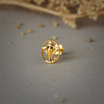 A gold oval stud earring with a Ganesha silhouette design, placed on a grey background with dried flowers