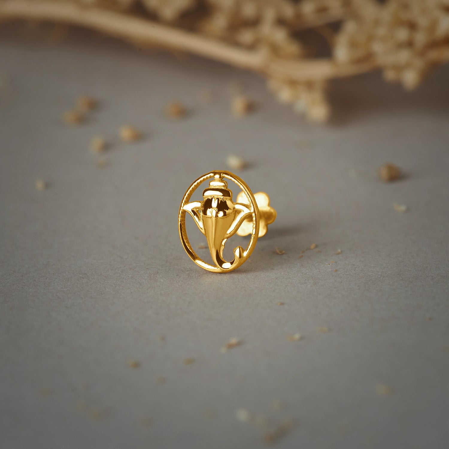 A gold oval stud earring with a Ganesha silhouette design, placed on a grey background with dried flowers