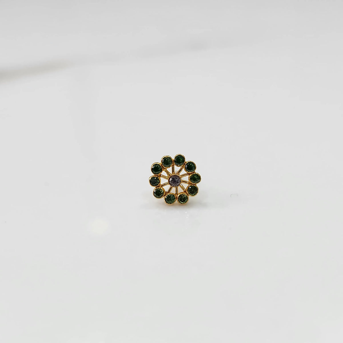 Front view of large gold nose pin with green gemstone petals and purple center stone on white background
