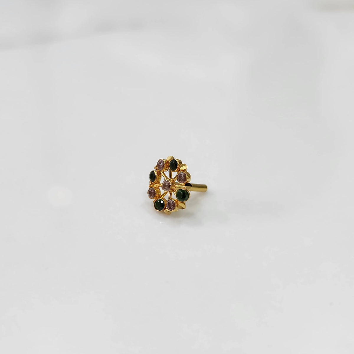 Gold pin nose with compact floral diamond nose pin set with alternating green and purple stones around a white center stone, placed on a white background