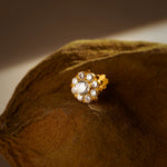 A gold stud earring with a large white center stone surrounded by a halo of smaller white stones, placed on a textured brown fabric background