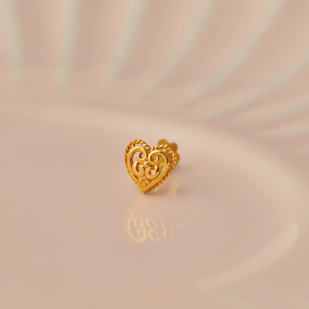 Gold heart-shaped nose stud with filigree scrollwork and twisted border on beige background