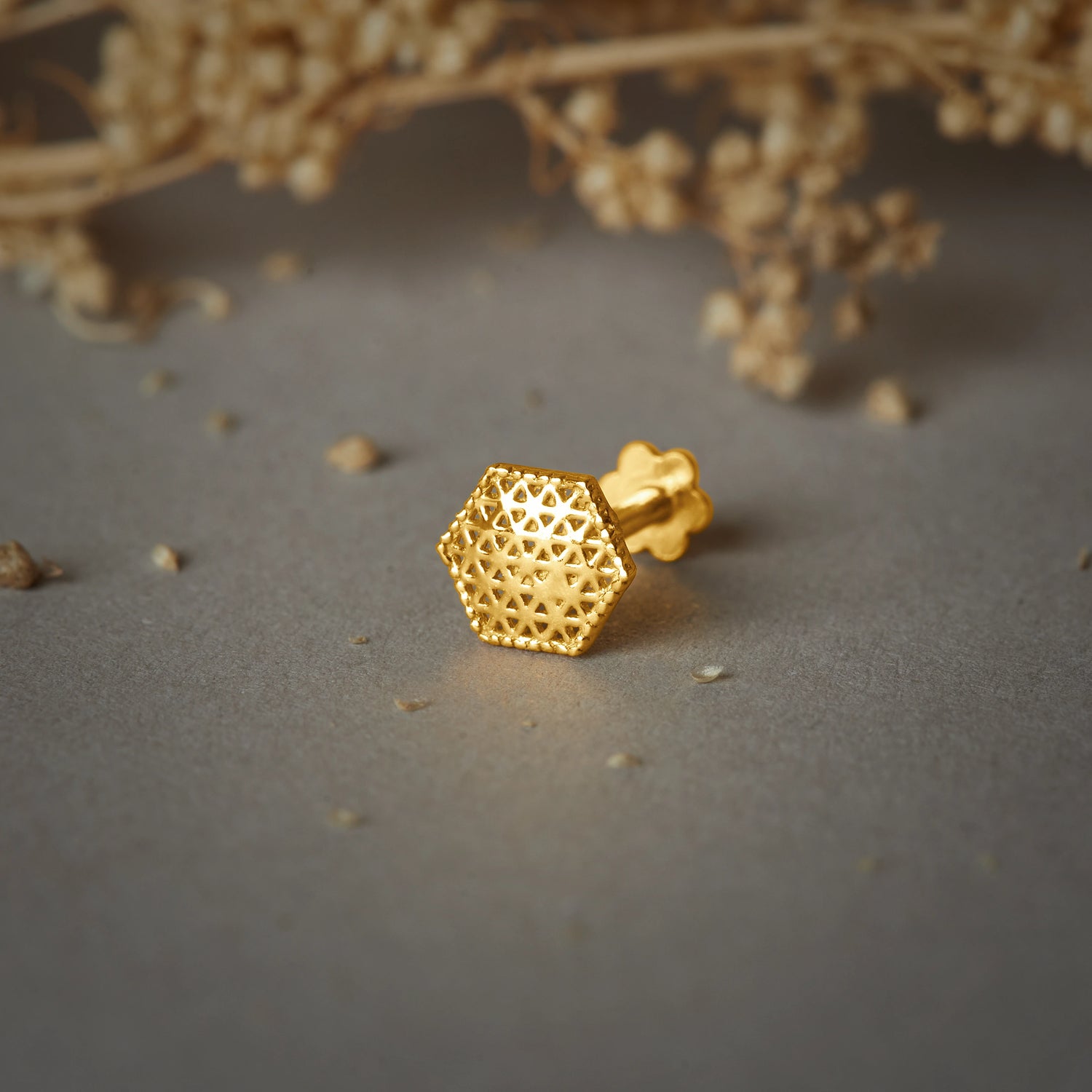 A gold hexagon-shaped stud earring with a jali cutwork pattern, placed on a grey background