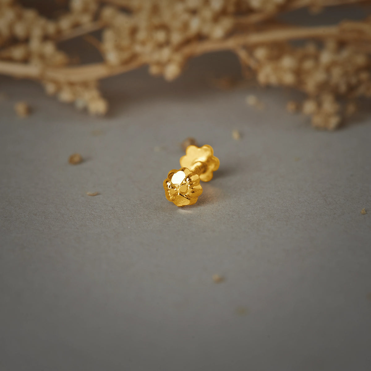A small gold flower-shaped stud earring with a hammered texture, placed on a grey background