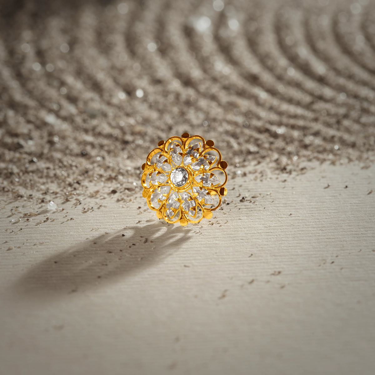A gold layered-petal stud earring with a center white stone surrounded by small white stone accents, placed on a textured beige fabric background