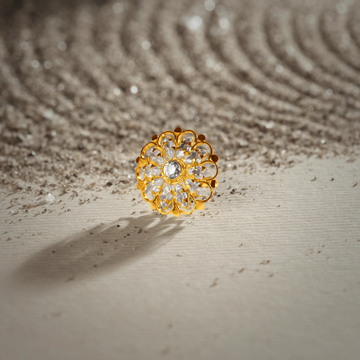 A gold layered-petal stud earring with a center white stone surrounded by small white stone accents, placed on a textured beige fabric background