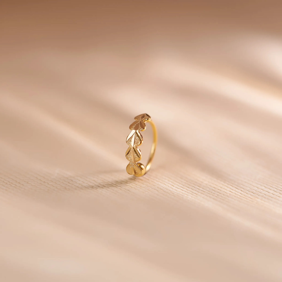 Close up of leaf patterned 18K gold nose ring standing upright on a soft beige surface with flowing light shadows