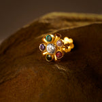A gold cluster stud earring with a white center stone surrounded by multicolor stones in teal, purple, pink, red and green, placed on a textured brown fabric background
