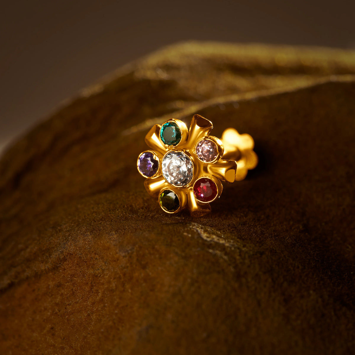 A gold cluster stud earring with a white center stone surrounded by multicolor stones in teal, purple, pink, red and green, placed on a textured brown fabric background