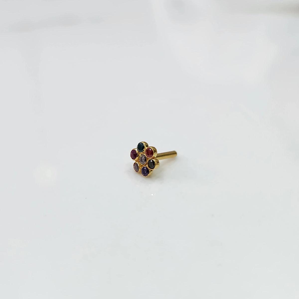 A gold flower-shaped nose pin with oval-cut multicolor stones in teal, red and purple, placed on a white background