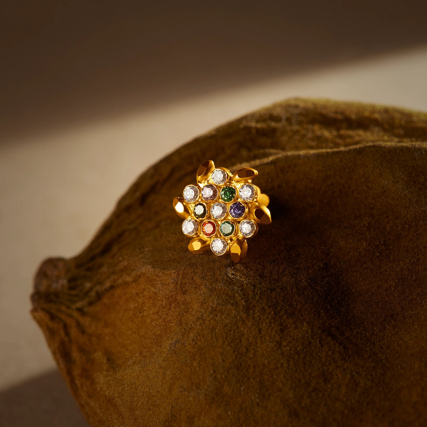 A gold stud earring with pointed petal edges and a cluster of multicolor stones in white, green, red and purple, placed on a textured brown fabric background