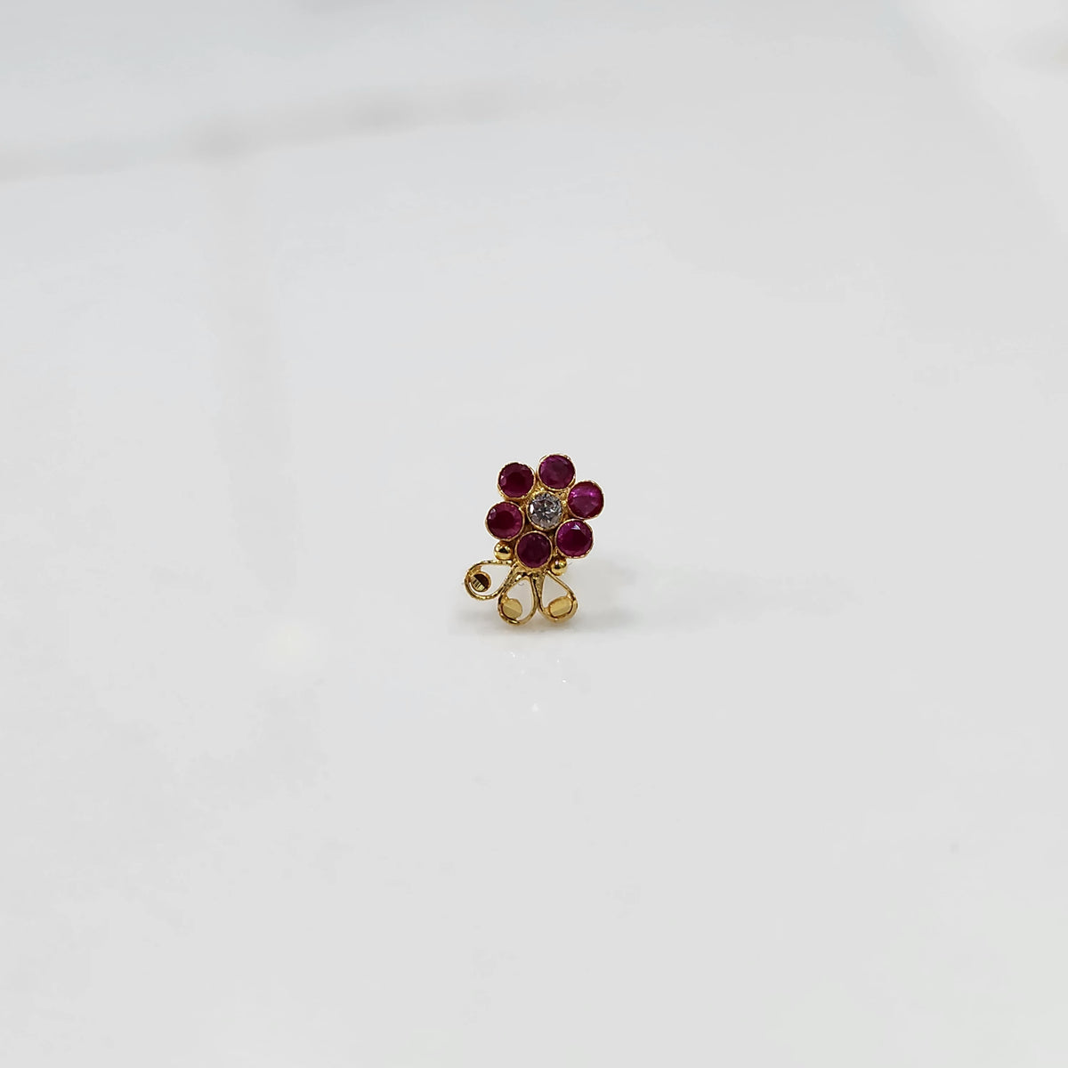 Traditional gold nose stud with ruby outer stones and clear center, decorative gold swirl below