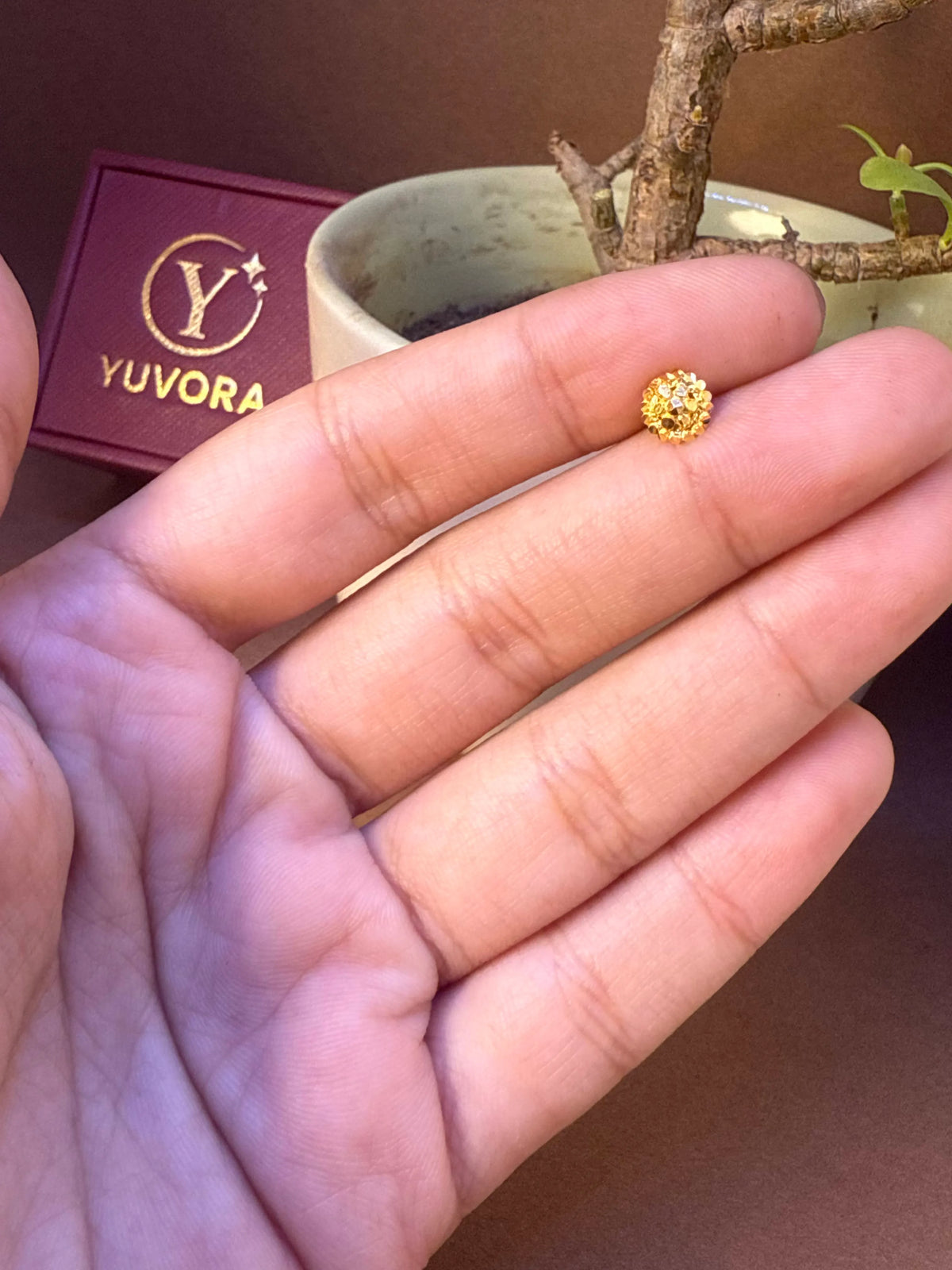 Round beaded gold nose pin with visible wire pin, held in hand, Yuvora jewelry box in background