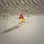 A gold oval drop-style stud earring with three ruby stones at the top and two white stones below, placed on a textured beige fabric background