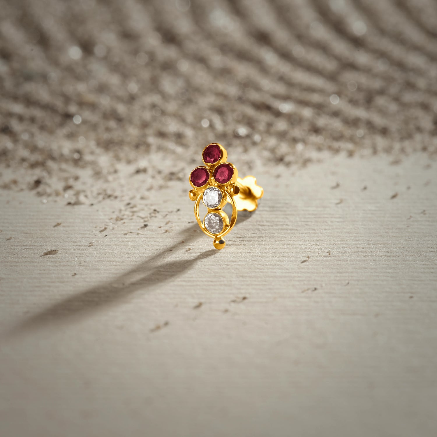 A gold oval drop-style stud earring with three ruby stones at the top and two white stones below, placed on a textured beige fabric background