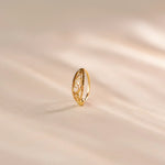 Close up of an elegant 18K gold nose ring with delicate openwork swirl detailing standing upright on a soft beige textured surface with gentle light shadows