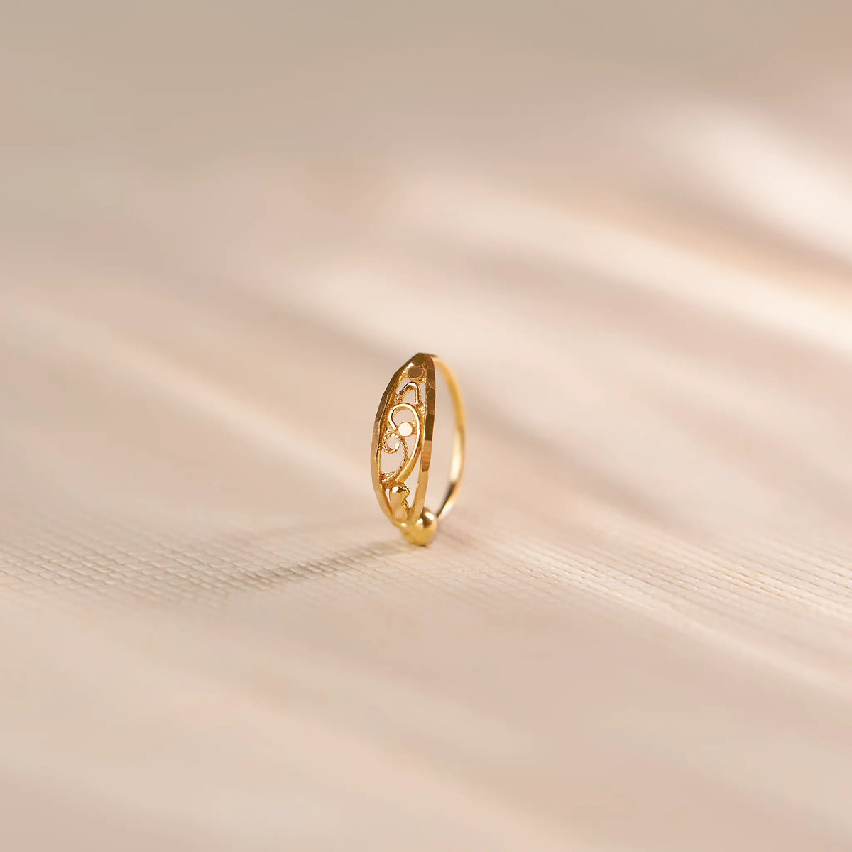 Close up of an elegant 18K gold nose ring with delicate openwork swirl detailing standing upright on a soft beige textured surface with gentle light shadows