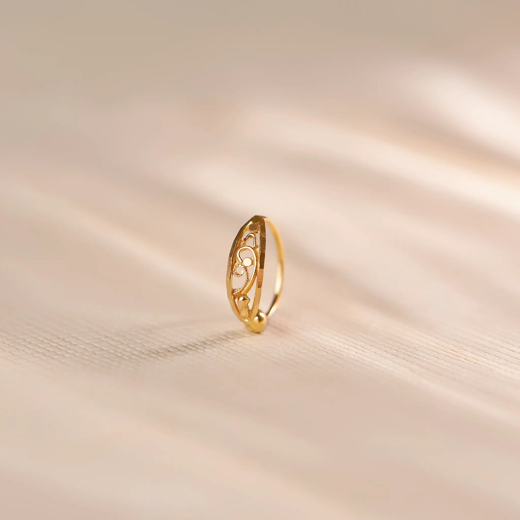 Close up of an elegant 18K gold nose ring with delicate openwork swirl detailing standing upright on a soft beige textured surface with gentle light shadows