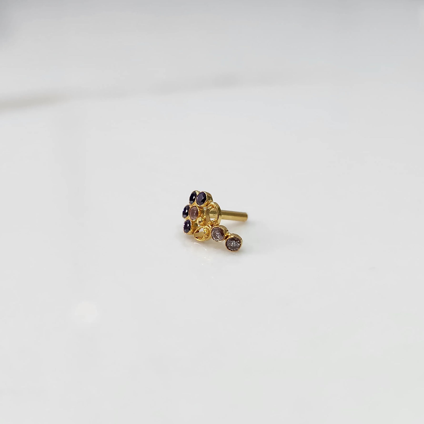 Front view of gold nose pin with purple gemstone cluster and clear drop-style stone accent on white background