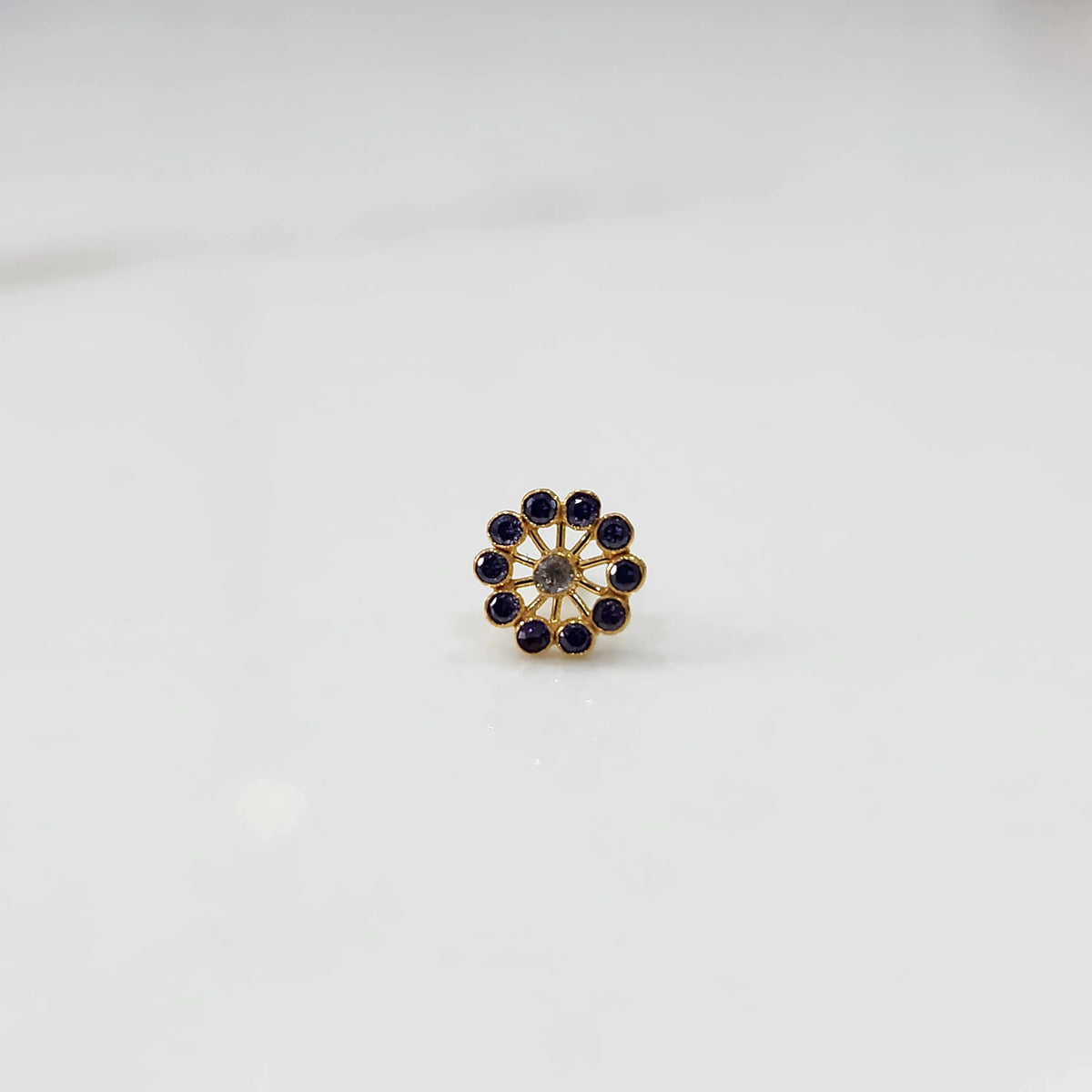 Front view of large gold nose pin with purple gemstone petals and clear center stone on white background