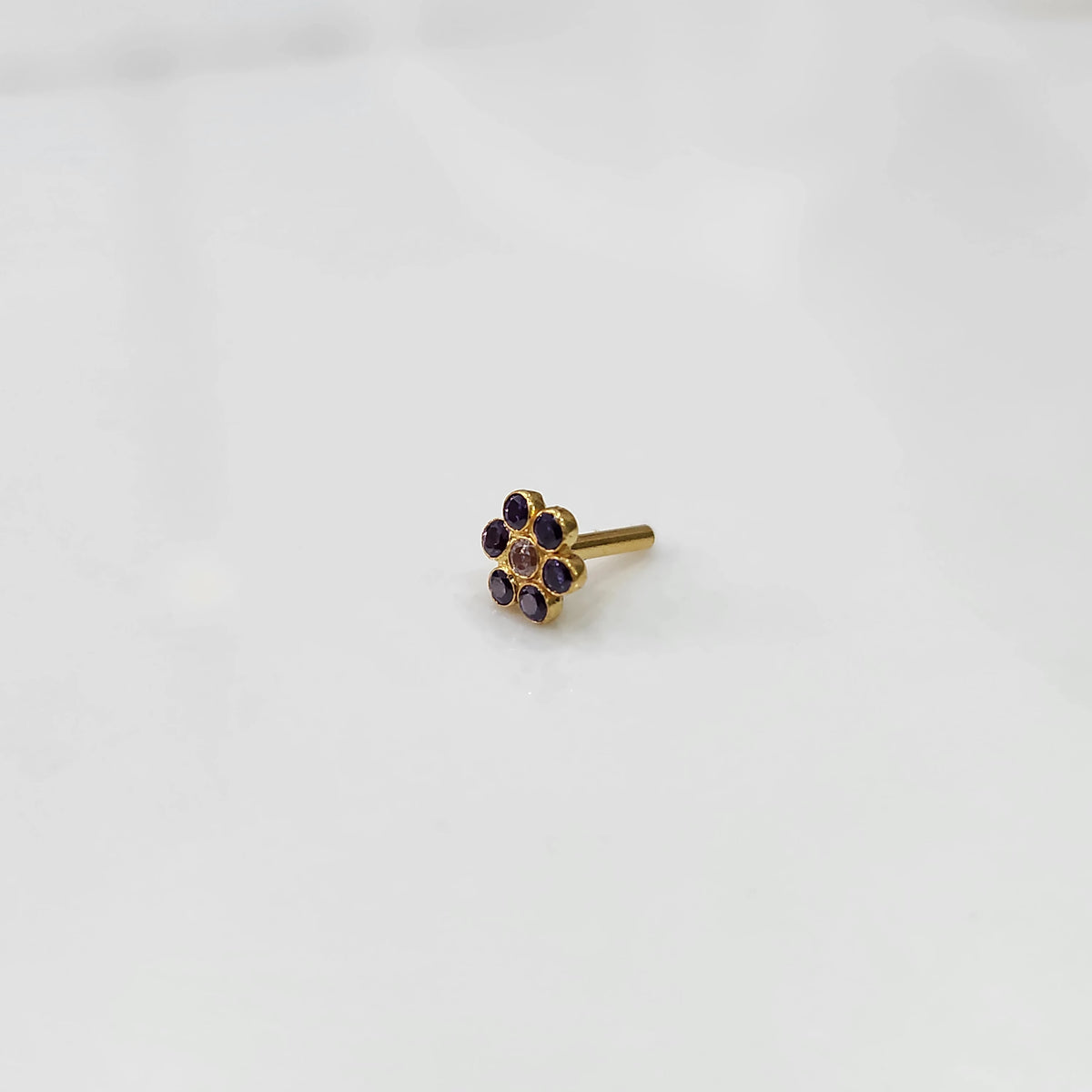A gold flower-shaped nose pin with purple stones around a small white center stone, placed on a white background