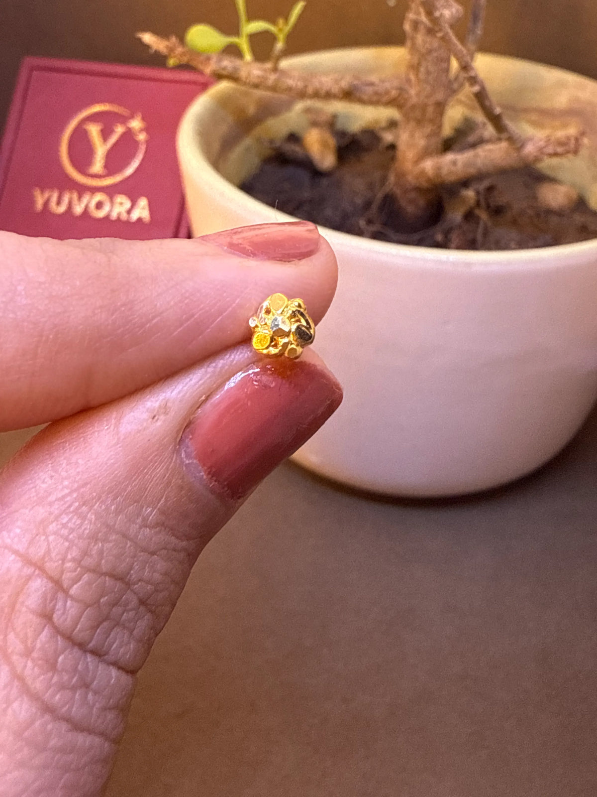 Small gold rosette-shaped nose pin stud held on fingertip near Yuvora jewelry box
