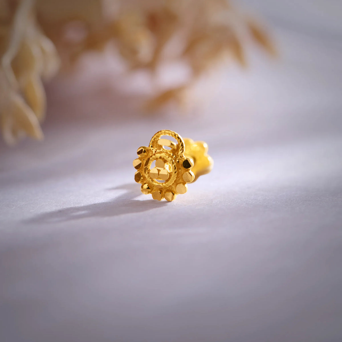 Close-up of a round gold nose pin with beaded border and screw-back fitting on a soft background
