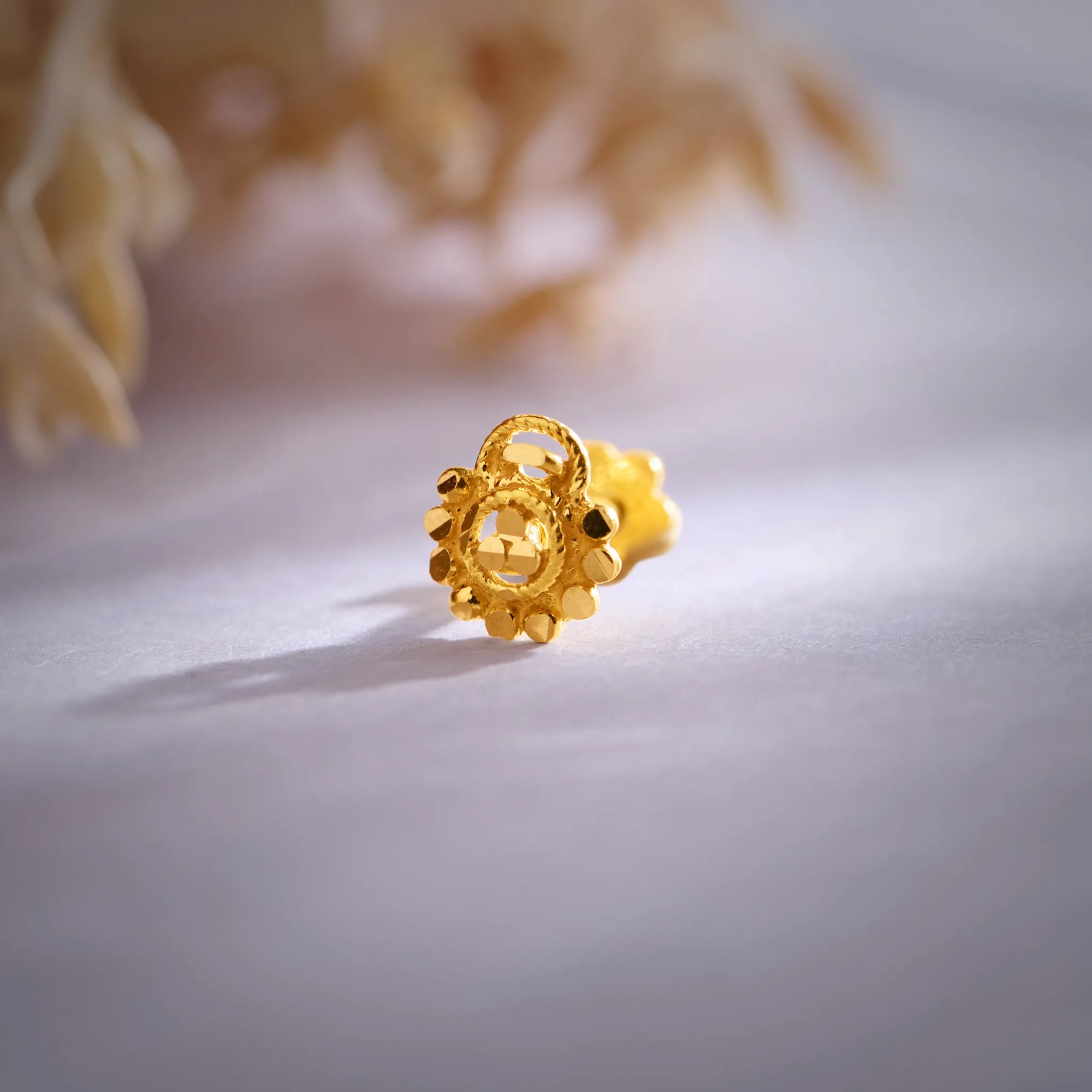 Close-up of a round gold nose pin with beaded border and screw-back fitting on a soft background